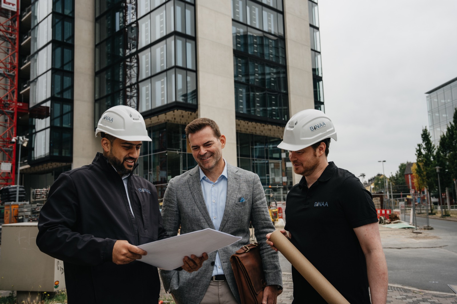 Drei laechelnde Maenner mit weissen Helmen betrachten Plaene vor einem Bauprojekt
