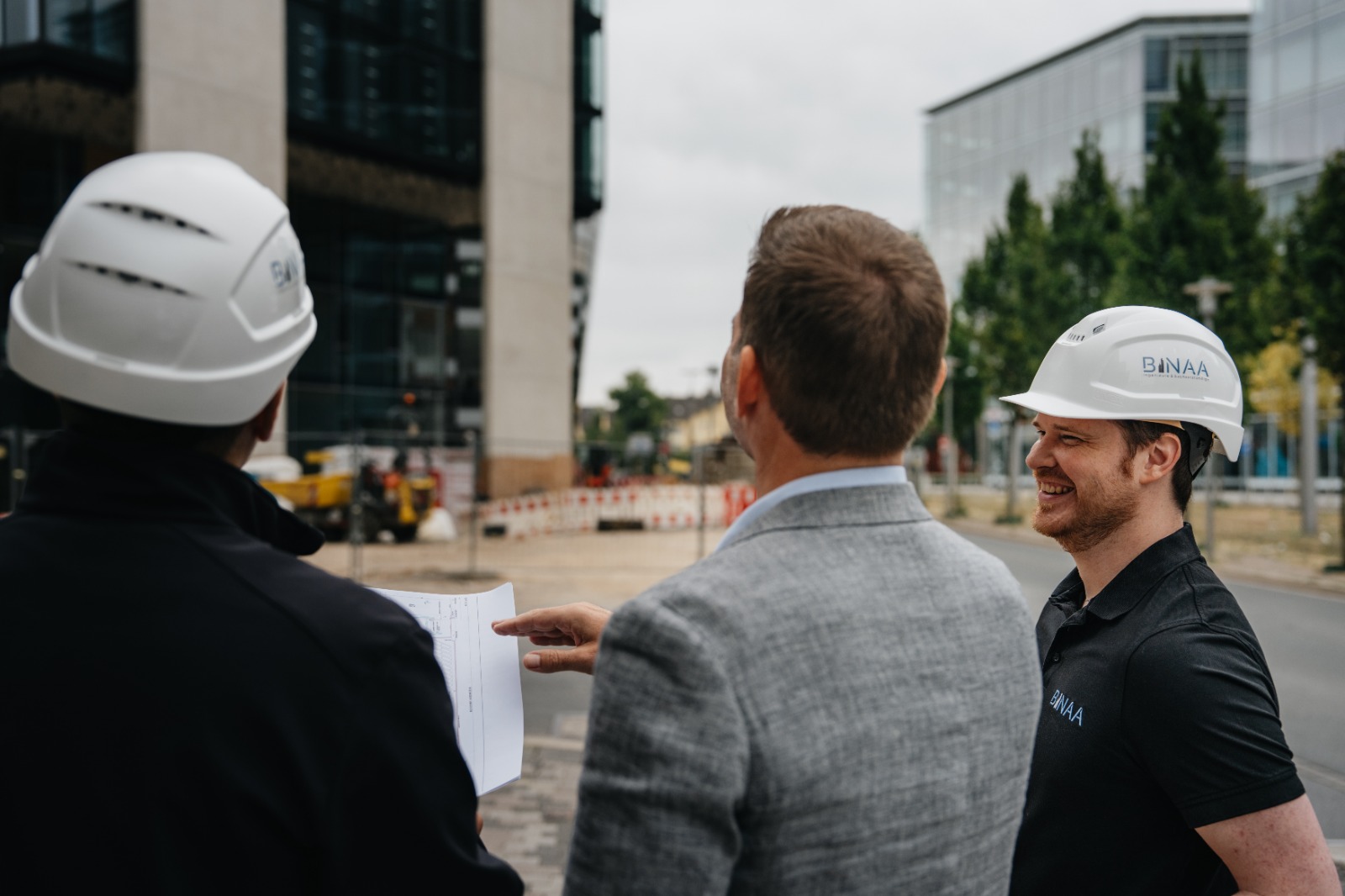 Drei Personen mit weissen Helmen besprechen laechelnd Baithemen auf der Baustelle