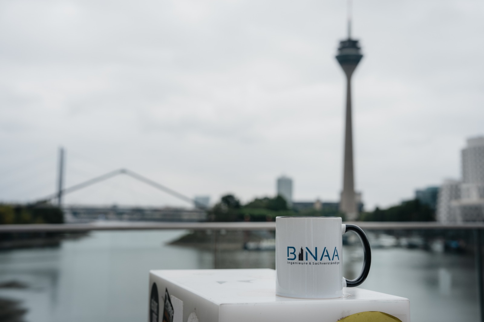 Weisse BINAA Tasse vor dem Duesseldorfer Rheinturm am bewoelkten Himmel